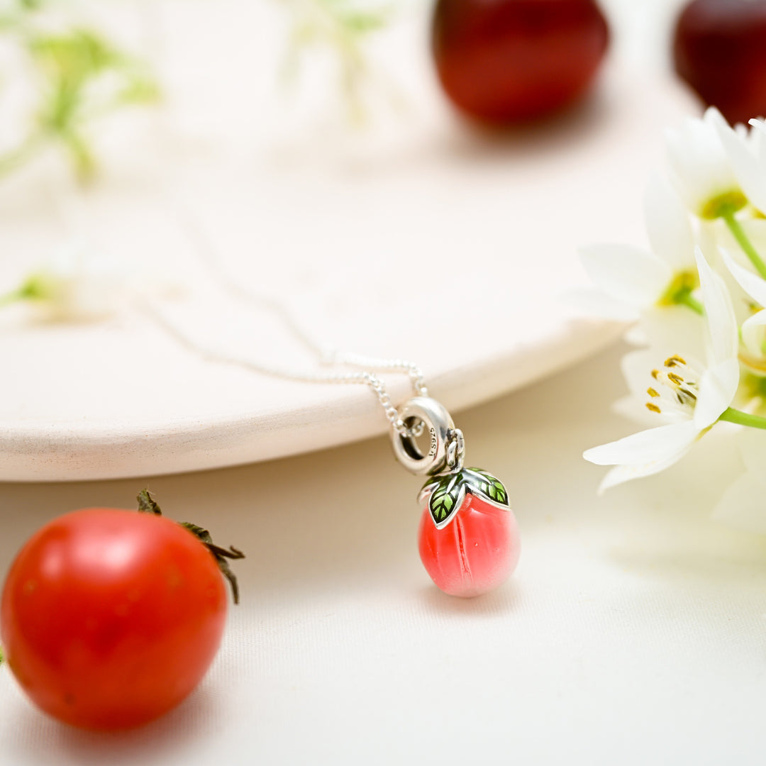Pink peach-shaped pendant necklace with silver loop and green leaves displayed among flowers and tomatoes