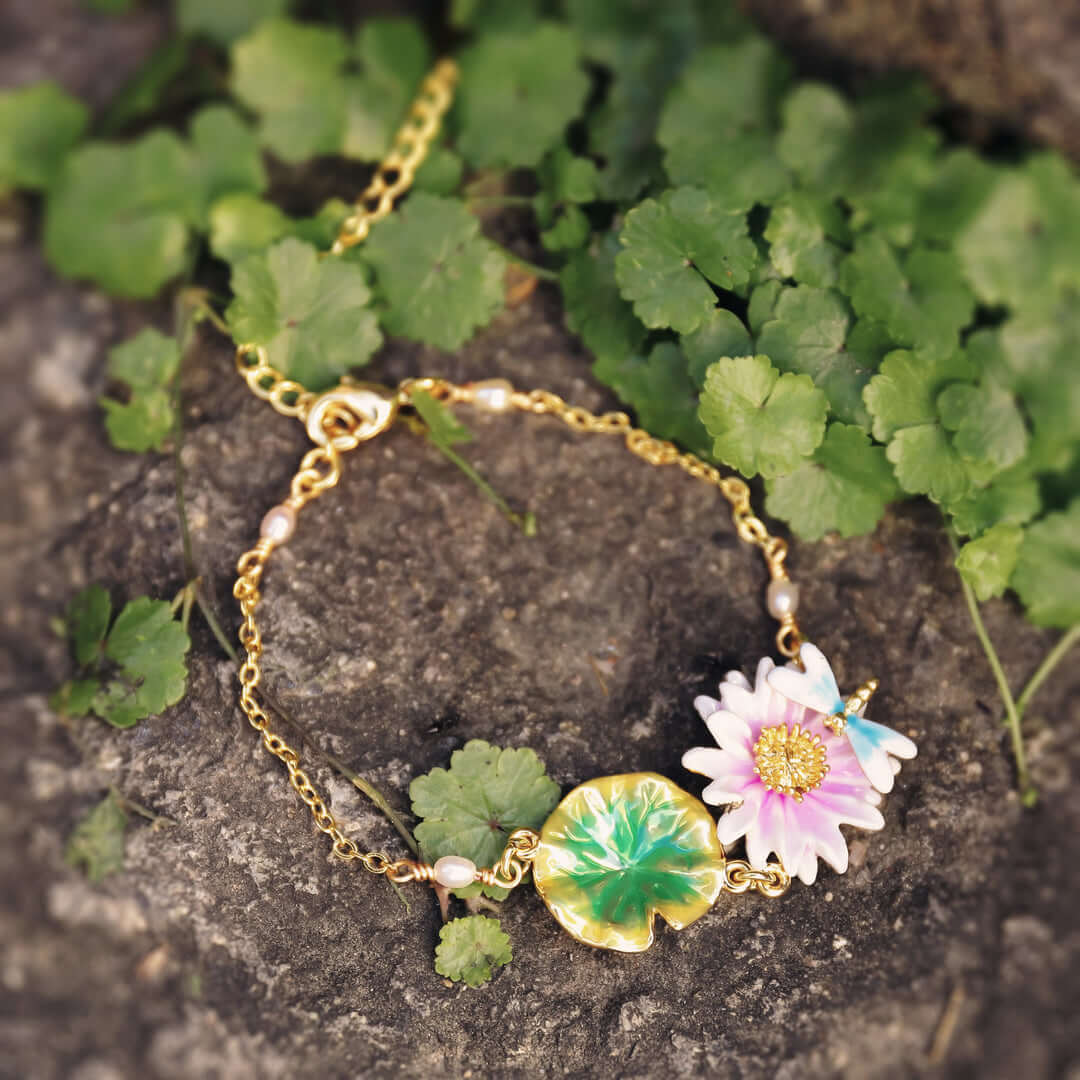 Nature-themed bracelet styled on dark stone with green plants, showing lotus flower, dragonfly charm, and gold chain.
