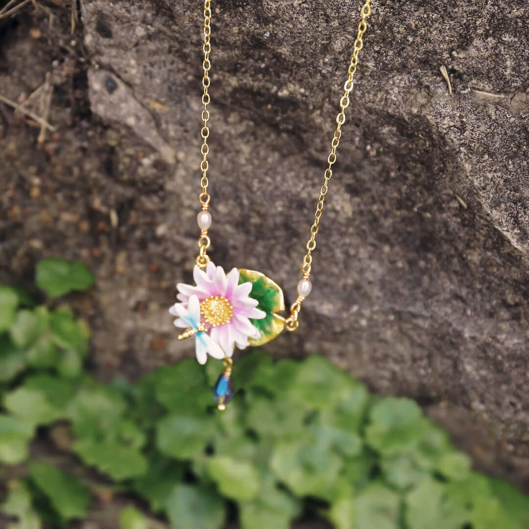 Lotus dragonfly necklace displayed on stone with soft greenery, featuring gold chain and pink lotus enamel pendant.