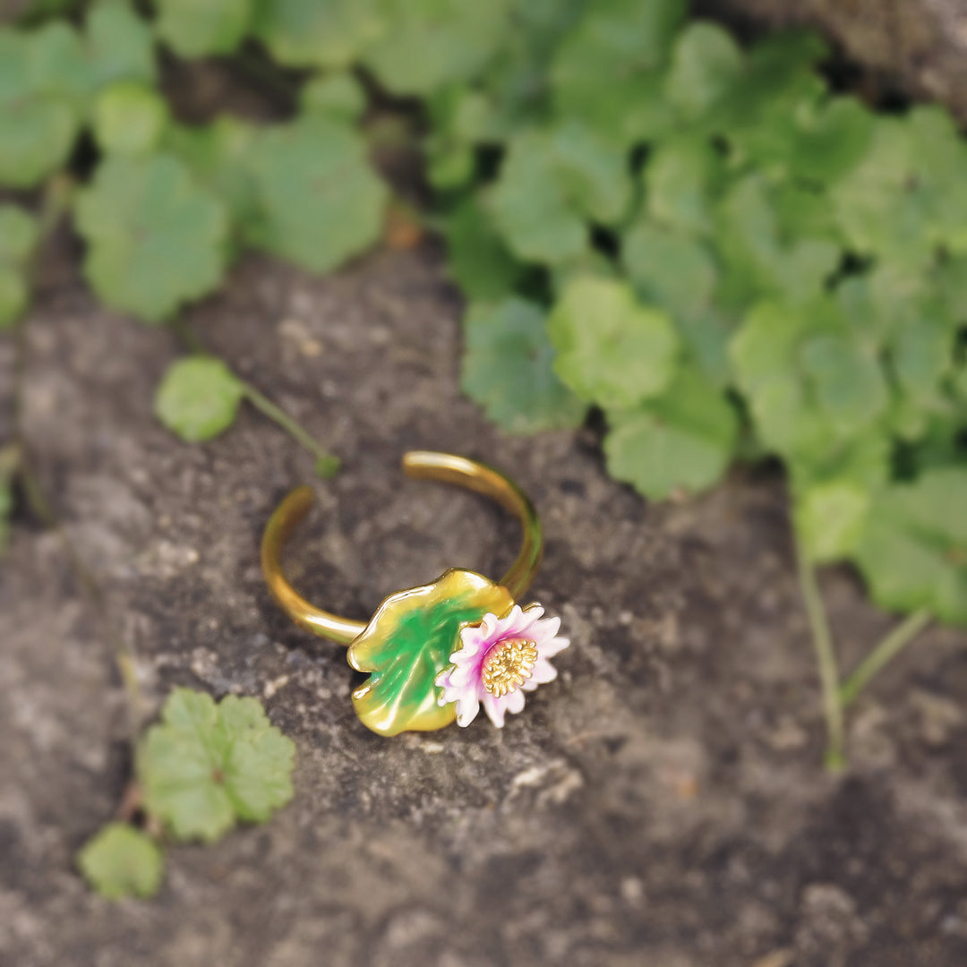 Lotus flower ring photographed on stone ground with green foliage, highlighting pink and green enamel.