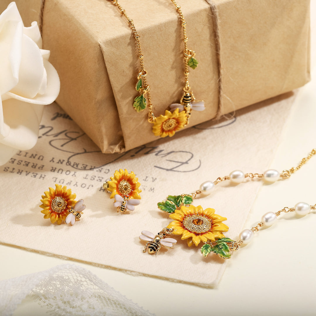 Flat lay of sunflower necklace and earrings set, featuring gold-tone bee charms, green leaves, and pearls next to a wrapped gift box