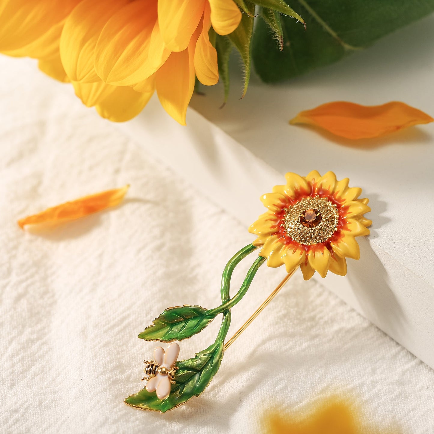 Sunflower brooch with enamel petals, green leaves, and a bee charm, displayed with sunflower petals on white cloth