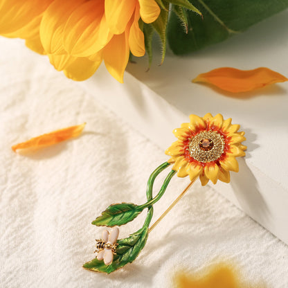 Sunflower brooch with enamel petals, green leaves, and a bee charm, displayed with sunflower petals on white cloth
